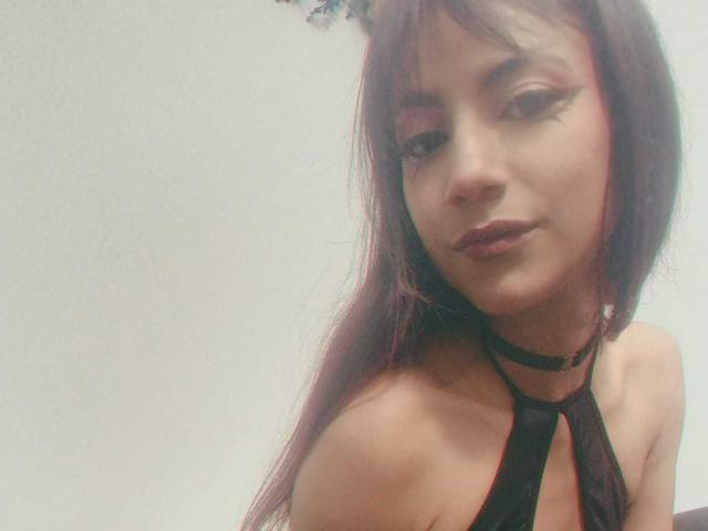 KarlaMiilan mit langem, violettem Haar, schwarzem Eyeliner und dunklem Lippenstift, trägt ein schwarzes Top mit Halter, das ihre Schultern freilegt, und eine Halskette. Sie hat ein leichtes, selbstbewusstes Lächeln..