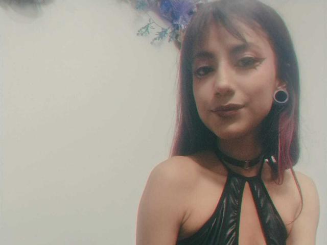 KarlaMiilan with purple-dyed hair, black leather halter top, choker, and gauges, smiling slightly, showing dark lipstick and winged eyeliner. Her small tits are partially visible.