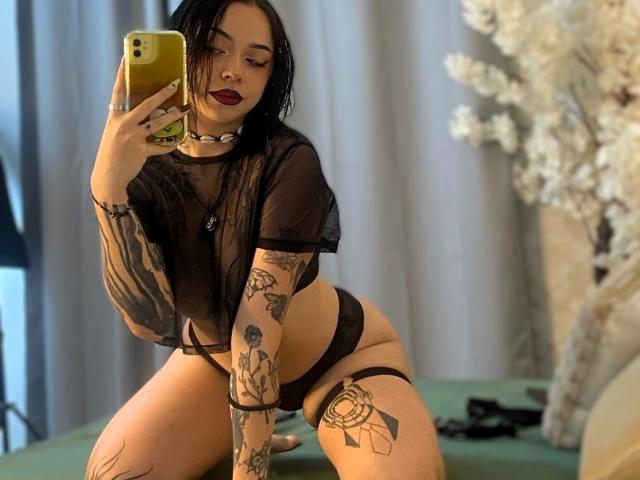 NaiaWhisper with black hair, tattoos, and red lipstick, wearing a sheer black top and black panties, kneeling on a green bed, holding a yellow phone, showing her tits.