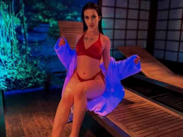 IsabelleDelicious sits on a wooden bench in a dimly lit room. She wears a red bikini with a strappy design, and an open blue robe. Her long brown hair is slicked back, and she has a tattoo across her chest. Her legs are crossed, and she holds the robe's edges. The lighting is blue and green, highlighting her toned body and fair skin. The bench is wooden, and the background includes a shoji screen. The image has a vivid, almost digital quality.
