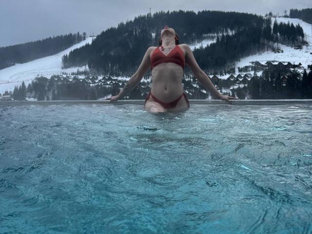 IsabelleDelicious stands in a pool, water reaching just below her red bikini. She's centered with arms out, eyes closed. Her red hair is tied back, and she has a fair skin tone. She's slightly overweight with a soft belly. The pool water is clear with subtle ripples. Snow-covered hills and forested slopes are in the background. The image is a realistic photograph with natural light, showing a winter scene. The overall tone is cool with blue water and white snow.