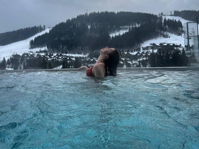 IsabelleDelicious, a woman with wet dark hair, is floating in a blue-tiled outdoor pool, her head tilted back, eyes closed. She's wearing a red one-piece swimsuit. Her upper body is visible, with water ripples around her. The background shows a snowy mountain with dark green trees and a small, dark-roofed building. The image has a cool, overcast tone, with grey skies and muted colors. Water texture is clear, with light reflections. IsabelleDelicious is centered, with the pool's edge slightly out of frame.