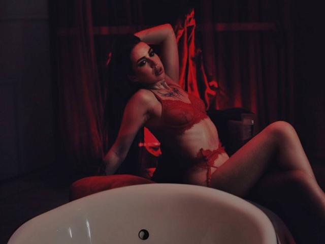 IsabelleDelicious sits in a dark room with red lighting. She wears red lace lingerie, her right arm raised, left hand on her thigh. Her long dark hair is slightly messy. She has a tattoo on her chest. Her legs are spread, one knee bent. A white sink is in the foreground. The red light casts shadows on her pale skin. Her expression is sultry. The image has a dark, seductive atmosphere.