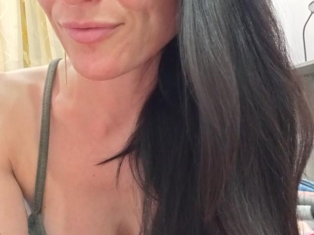 BellaJulls with long black hair, slight smile, and pink lips, showing upper chest and green strap, with visible cleavage and tan skin.