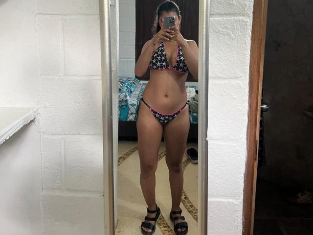 AdeFranco in a black and pink floral bikini, standing in front of a mirror, holding a phone, with visible tits, ass, and pussy, wearing black sandals, mirror reflection.