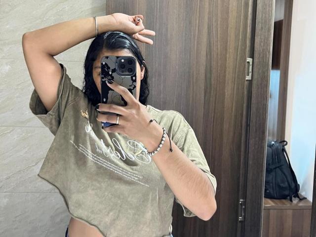 AdeFranco with dark hair, wearing a green crop top, taking a mirror selfie with one arm raised. Boobs not visible, hand covering face, phone with black case.