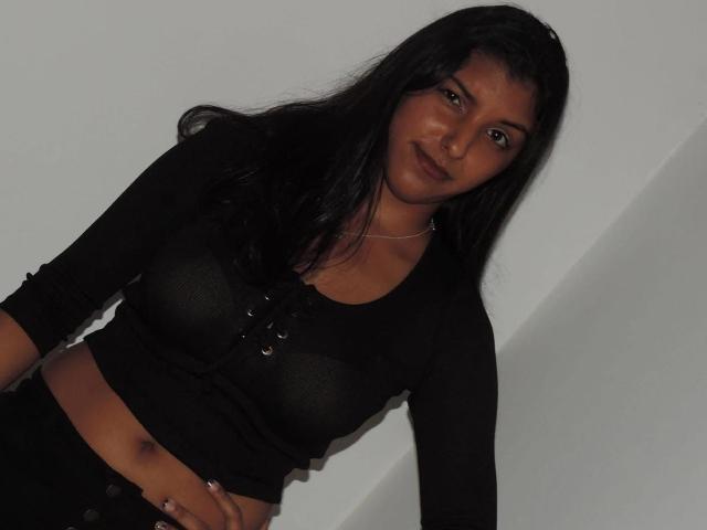 AdeFranco with long black hair, brown skin, wearing a black long-sleeve crop top, showing her small tits and midriff, standing against a white wall, looking at camera.