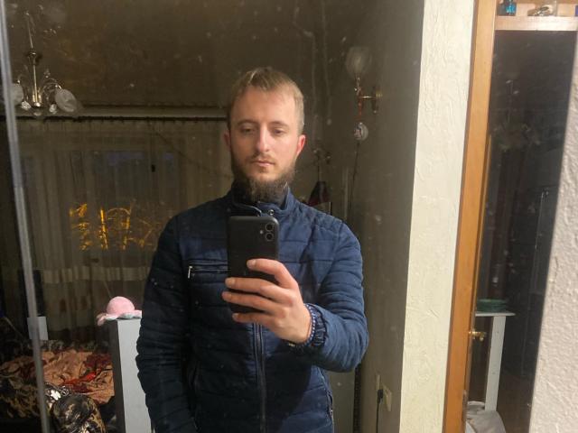DesireLarry, un homme barbu avec une courte coupe de cheveux blonde, portant une veste matelassée bleu marine, se prend en photo dans un miroir avec un téléphone noir..