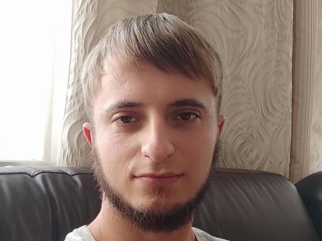 Larry hat kurze, braune Haare, einen leichten Bart und ein leichtes Lächeln, braune Augen und trägt ein weißes Hemd. Er sitzt auf einer dunklen Ledercouch..