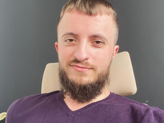 Larry tiene una barba espesa y oscura, cabello castaño corto, y lleva una camiseta morada con cuello en V. Está sonriendo ligeramente, con una expresión relajada y segura..