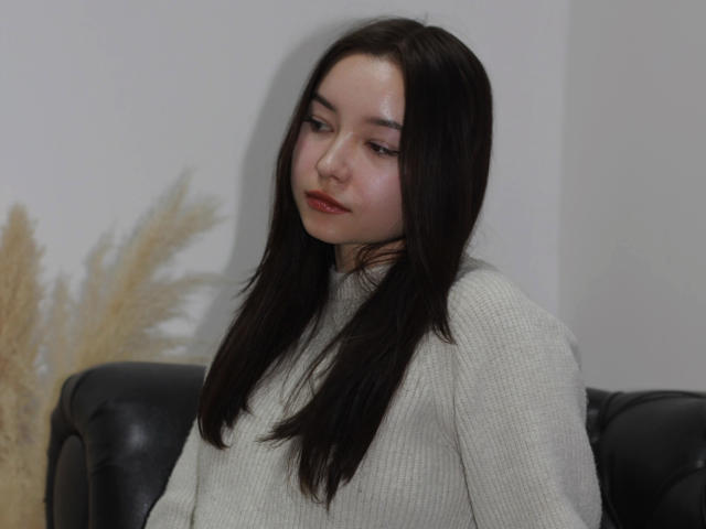 StellaBarnes with long black hair, pale skin, and red lipstick, wearing a white textured sweater, sitting on a black leather couch, looking down with a slight, relaxed expression.