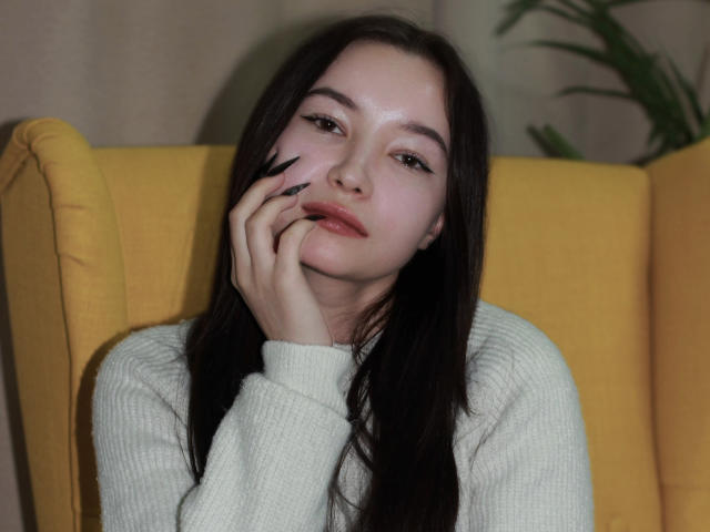 StellaBarnes with long black hair, pale skin, and dark eyes, wearing a white knit sweater, touching her face with black-manicured fingers, sitting on a yellow chair.