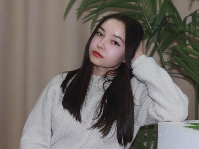 StellaBarnes with long dark hair, pale skin, and red lips, wearing a white knit sweater, hand in hair, green plant in background, looking slightly to the side.