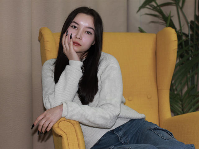 StellaBarnes with long dark hair, pale skin, and black nails, wearing a white sweater and blue jeans, sits in a yellow chair, hand on cheek, looking at camera.