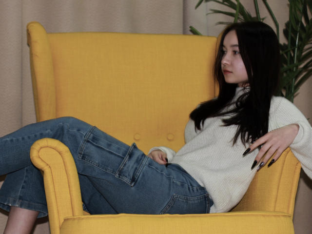 StellaBarnes with long black hair, wearing a white sweater and blue jeans, lounging on a yellow armchair, looking to the side with a relaxed expression.