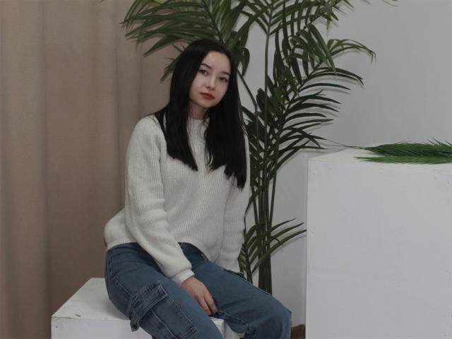 StellaBarnes with black hair, wearing a white sweater and blue jeans, sitting on a white cube, hands on her knee, green plant behind her, beige curtain on left.