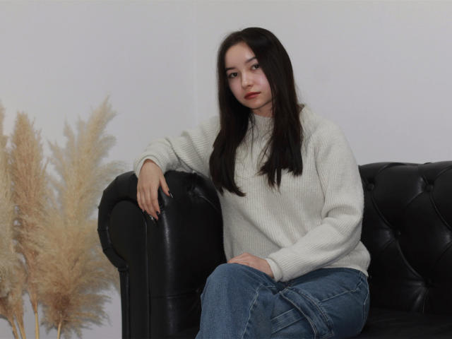 StellaBarnes with long black hair, white sweater, and blue jeans, sitting on a black leather couch, arm resting on the armrest, looking at the camera, puffy pants.