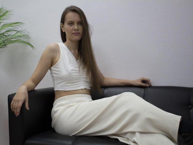 AyumiKirk with long brown hair, wearing a white crop top and white skirt, sitting on a black leather couch, showing her slim arms and small tits.