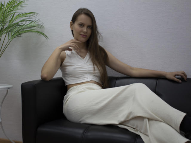 AyumiKirk with long brown hair, wearing a white crop top and white pants, sitting on a black leather couch, legs crossed, right hand near chin, black socks.