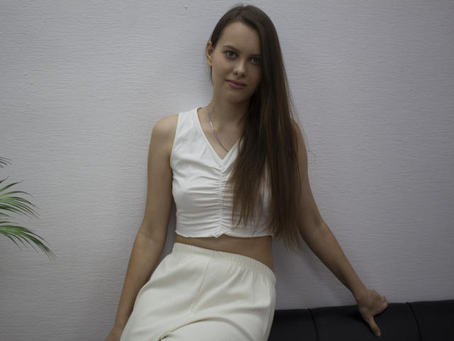 AyumiKirk with long brown hair, small tits, wearing a white crop top and high-waisted white pants, sitting on a black couch, smiling slightly.