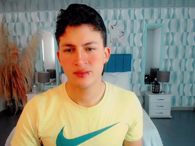 AxelSavage a les cheveux noirs et bouclés, courts, un petit piercing au nez et une coupure sur la lèvre inférieure. Il porte un t-shirt jaune Nike avec un logo vert en forme de virgule..