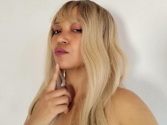 SuperYara, con cabello rubio, párpados rosados y lápiz labial rosa, tocándose la barbilla con un dedo, sonriendo sutilmente. Tiene piel clara y un aspecto lindo y sexy..