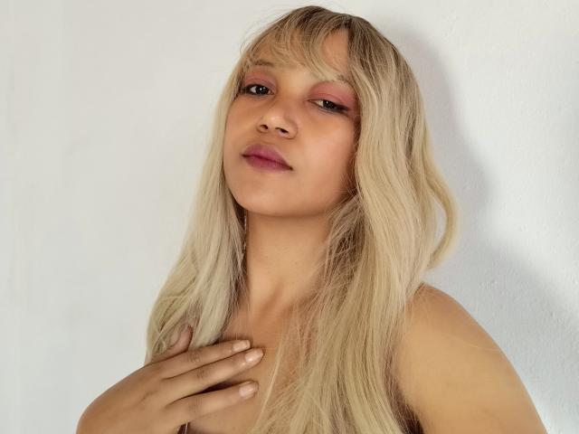 SuperYara com cabelo loiro, sombra rosa nos olhos e lábios nude, com a mão no peito, olhando para a câmera, sorriso suave, fundo de parede branco. Sem seios aparentes, sem bumbum visível, sem vulva visível..