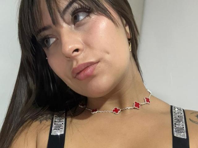 QuennAfrodita with dark brown hair, blue eyes, and a slight pout. She's wearing a red flower necklace and a black, sparkly top, showing a small tattoo on her right shoulder.
