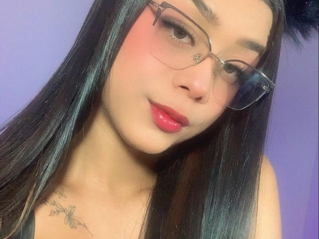 Elyw has long black hair, red lipstick, and glasses, with a small tattoo on her left chest. Her pink cheeks and subtle smile are noticeable.