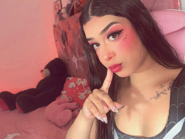 Elyw with pink eyeliner and red blush, finger in mouth, long dark hair, tattoos on chest, pink lips, black nails, in pink-lit room, black teddy bear in background.