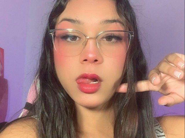 Elyw has pink makeup on her cheeks, red lipstick, and is wearing clear glasses. She's pointing at her face with her right hand, lips slightly parted.