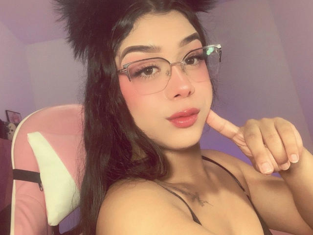 Elyw with black cat ears, glasses, pink blush, and pink lips, showing off her tits with a finger near her face, in a pink chair.
