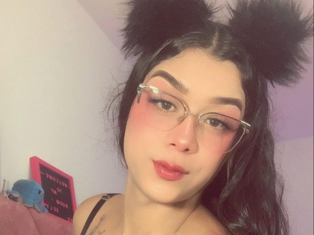 Elyw has black fluffy cat ears, glasses, pink blush, red lips, and black wavy hair, showing off her puffy tits in a selfie with a cute blue stuffed toy.