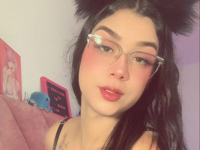 Elyw with black cat ears, glasses, pink blush, glossy red lips, and long black hair. Her tits are small, and she has a cute, sexy look.