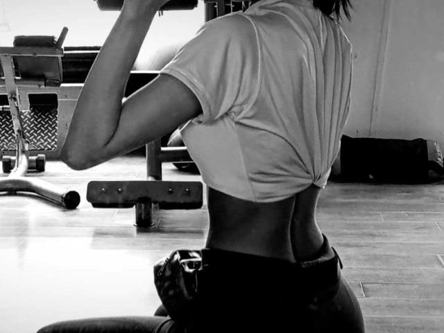 Black-and-white photo of DaphneSpencer with toned back, tied-up shirt, holding dumbbell, wearing dark pants, belt with buckle, and wooden floor. Her ass is visible.