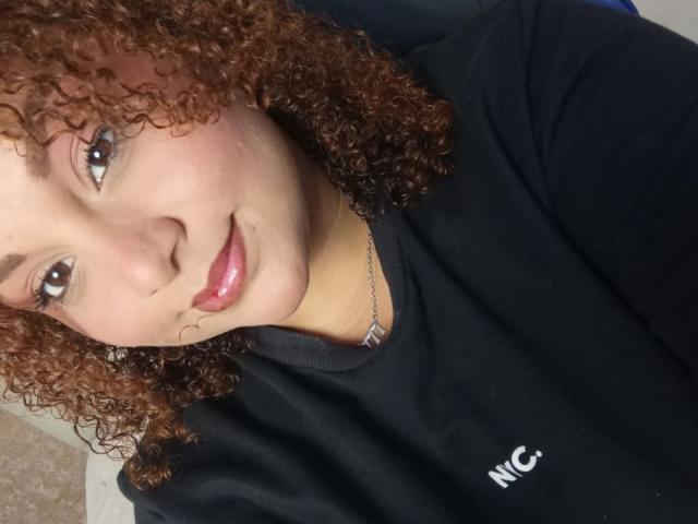YeraCarey with curly brown hair, light skin, and pink lips, wearing a black NC shirt, smiling softly at the camera. She's wearing a delicate silver necklace.