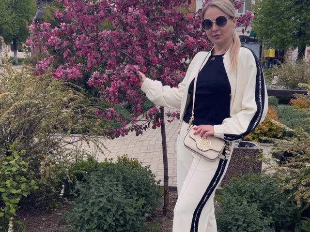 Zetrx69 with blonde hair, black top, white jacket with black stripes, white pants, black sunglasses, small white purse, pointing at pink flowering tree, standing in garden.
