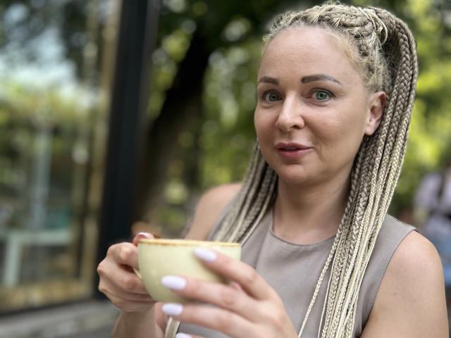 Zetrx69 with blonde braids, holding a yellow cup, wearing a sleeveless brown top, has pale pink lipstick and white nails, looking at the camera with a slight smile.