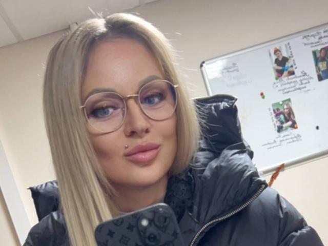 Zetrx69 has blonde hair, blue eyes, and wears round glasses with a nose piercing. She's in a black puffy jacket, looking at a phone with a pink case.