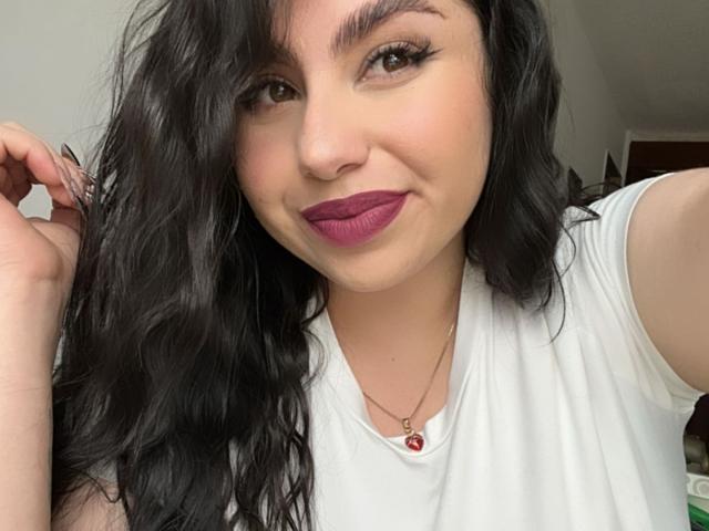 VenusxMusa with wavy black hair, wearing a white shirt, purple lipstick, and a necklace with a red gem, smiling at the camera, holding her hair with a finger.