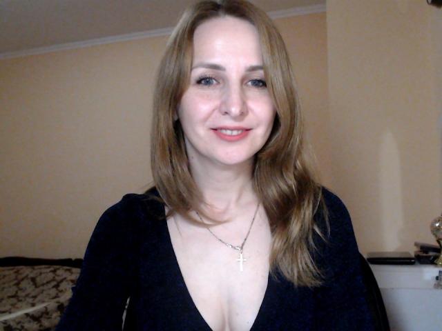 RuthPark, a white woman with long brown hair, smiling, wearing a black V-neck top, showing cleavage, and a cross necklace, in a dimly lit room.