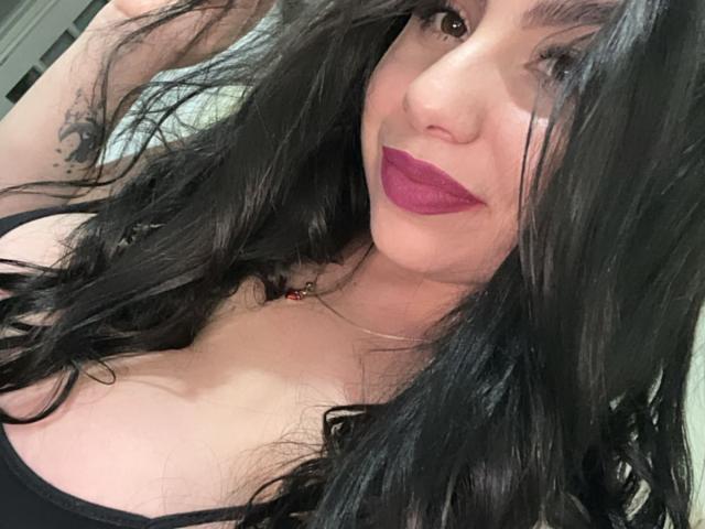 VenusxMusa mit langen schwarzen Haaren, lila Lippenstift und einem schwarzen Top, das ihre Brüste zeigt. Sie lächelt, hält ihr Haar und hat ein Tattoo am Arm..