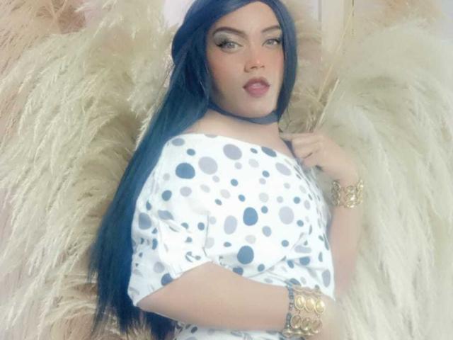 CharlotteSweetX with long blue hair, wearing a white polka dot dress, gold bracelet, and choker, lying on fluffy beige feathers, looking at the camera with full pink lips.