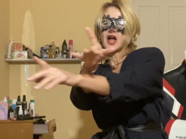 TianaQLove with blonde hair, black mask, and black dress, reaches out with hands, mouth open, in a bedroom with shelves and bottles. Her tits are partially visible.