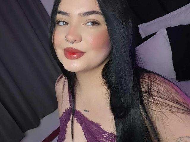ValeriJoness with long black hair, nose ring, and red lipstick, wearing a purple lace bra, showing a small tattoo under her chest, smiling at the camera.