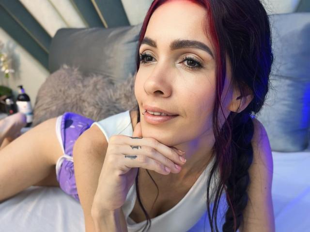 KathySteven, avec des cheveux roux et noirs, un petit anneau de nez, un débardeur blanc, un short violet, allongée sur un lit, un doigt sur le menton, un tatouage sur le doigt, souriante, éclairage bleu et rouge..
