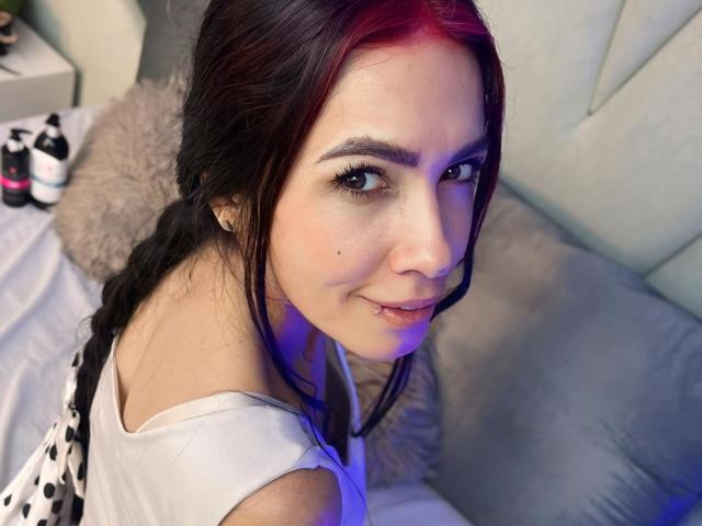 KathySteven with dark red highlights in her brown hair, smiling with a small lip piercing, wearing a white top, sitting on a bed with a fluffy pillow.