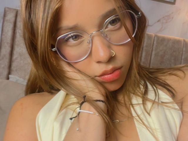 HeraJuno with glasses, nose ring, and pink lips, wearing a yellow top, touching her cheek with a hand, brown hair, and a bracelet, looking at the camera.