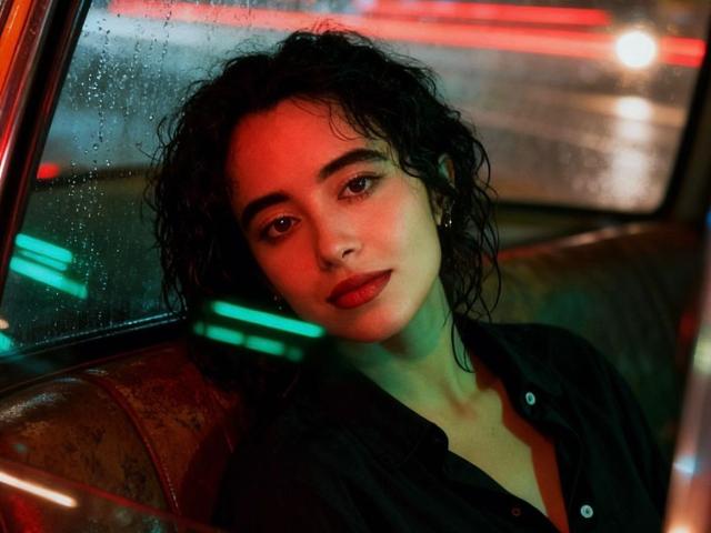 VivienneLaurent with curly black hair, red-lit face, and serious expression, wearing a black button-up shirt, leaning against a wet car seat, looking out the window.