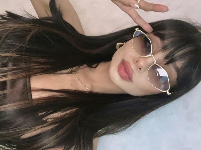 ThaliaCollins with long black hair, wearing gold sunglasses, a brown top, and a gold necklace, showing off her pouty pink lips and making a "rock on" hand gesture.